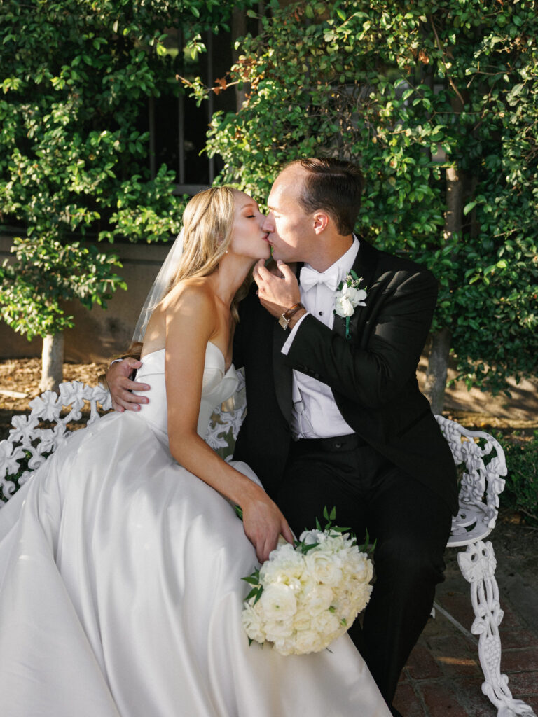 Romantic bride and groom kissing on an outdoor bench while the bride holds a classic lush bouquet of white garden roses