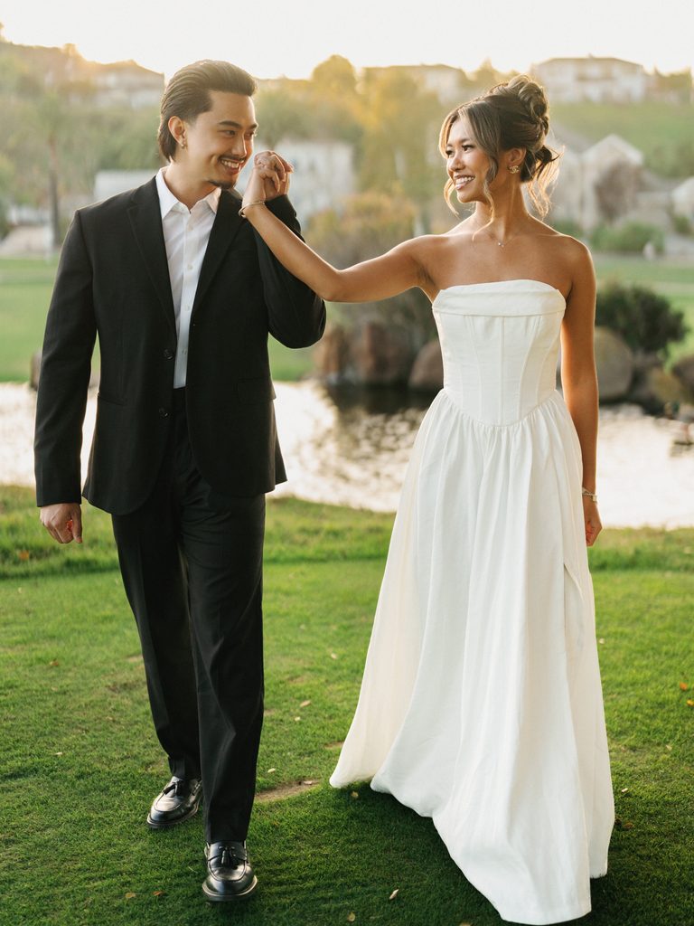 Chic newlyweds holding hands and walking on a green lawn near a scenic lake during a luxury golden hour portrait session
