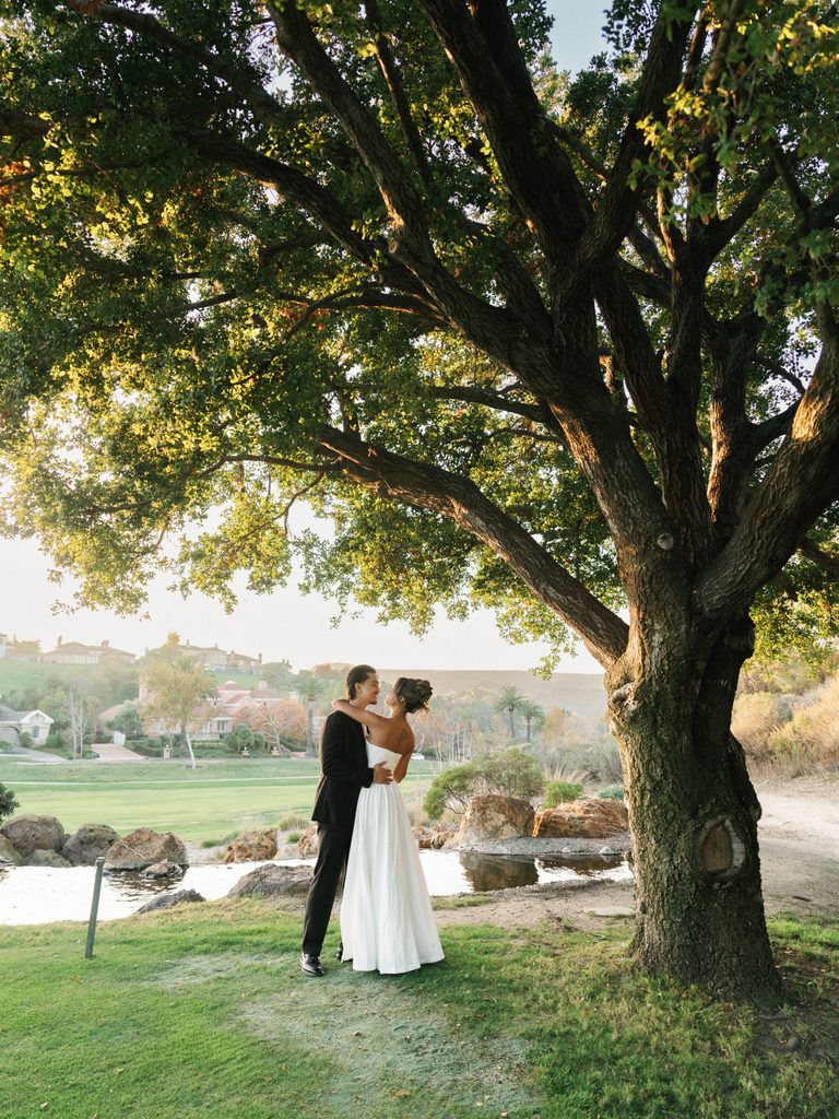 Cinematic wide shot of a couple embracing under a majestic old oak tree next to a scenic pond on a luxury estate golf course