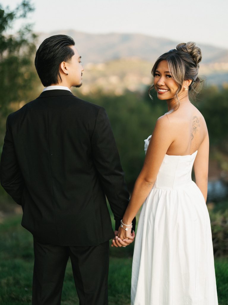 Beautiful bride looking back over her shoulder holding hands with her groom against a scenic mountain landscape background view