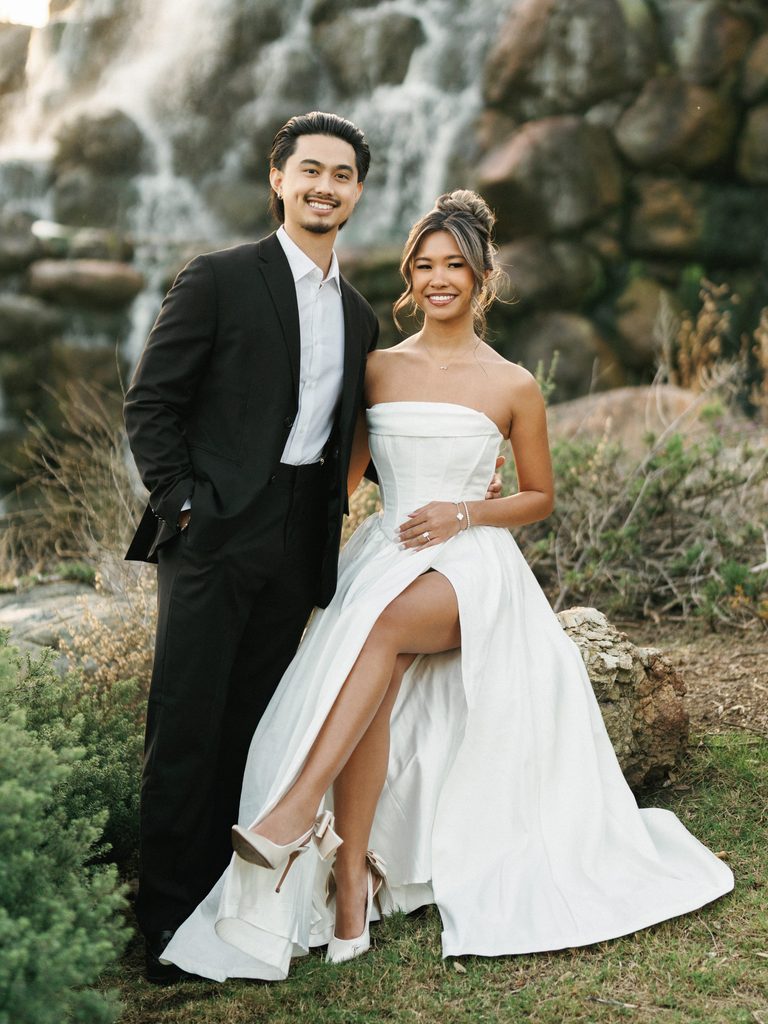 Stunning newlyweds posing outdoor in front of a waterfall background showcasing white bow bridal heels and a classic black groom suit