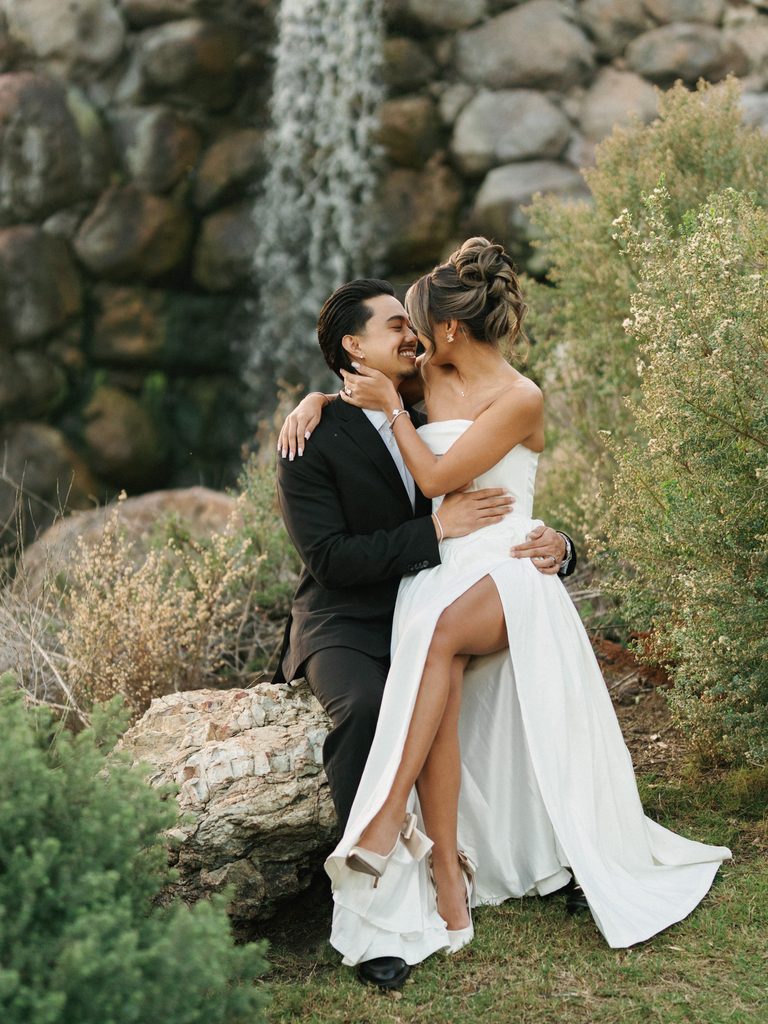 Playful bridal couple laughing together on a rock near a scenic waterfall featuring an elegant strapless gown with a high leg slit
