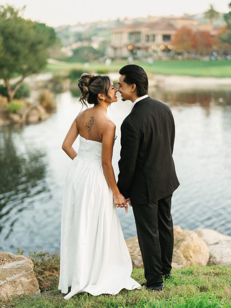 Intimate bridal couple standing on a grassy hill near a scenic lake looking at each other before a romantic kiss