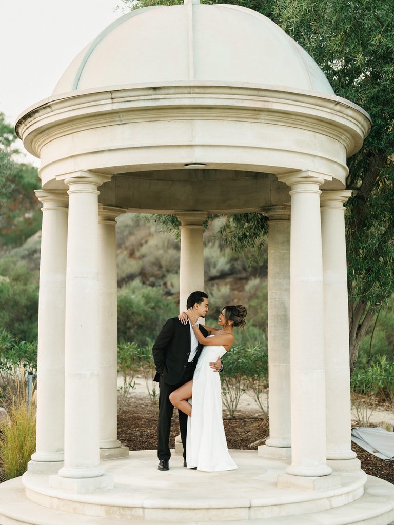 Cinematic wide shot of a couple embracing inside a majestic stone gazebo with classical columns surrounded by lush green nature