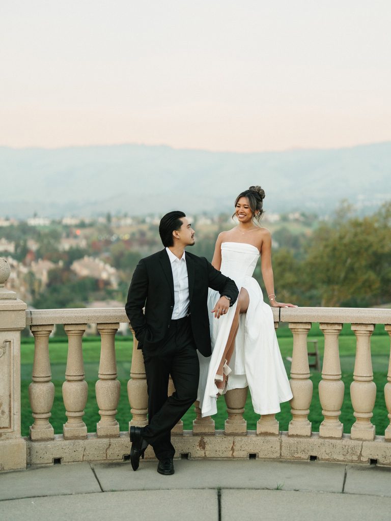 Modern bridal couple smiling and posing near a stone balustrade on an outdoor overlook terrace with a scenic mountain valley view