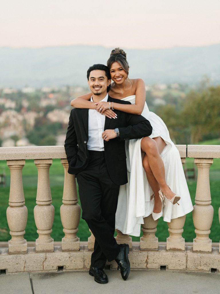 Happy bride sitting on a classic balcony rail and embracing her groom from behind during a luxury destination wedding photo session