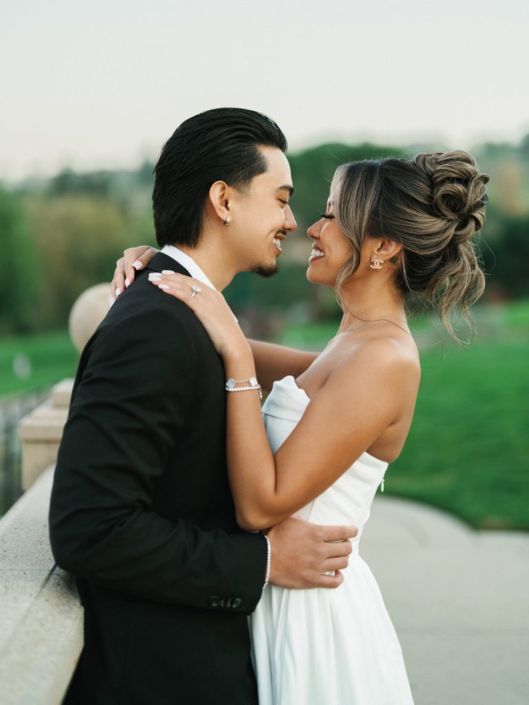 Elegant newlyweds smiling at each other showcasing a textured bridal corset a luxury clover charm bracelet and a stylish high bun updo
