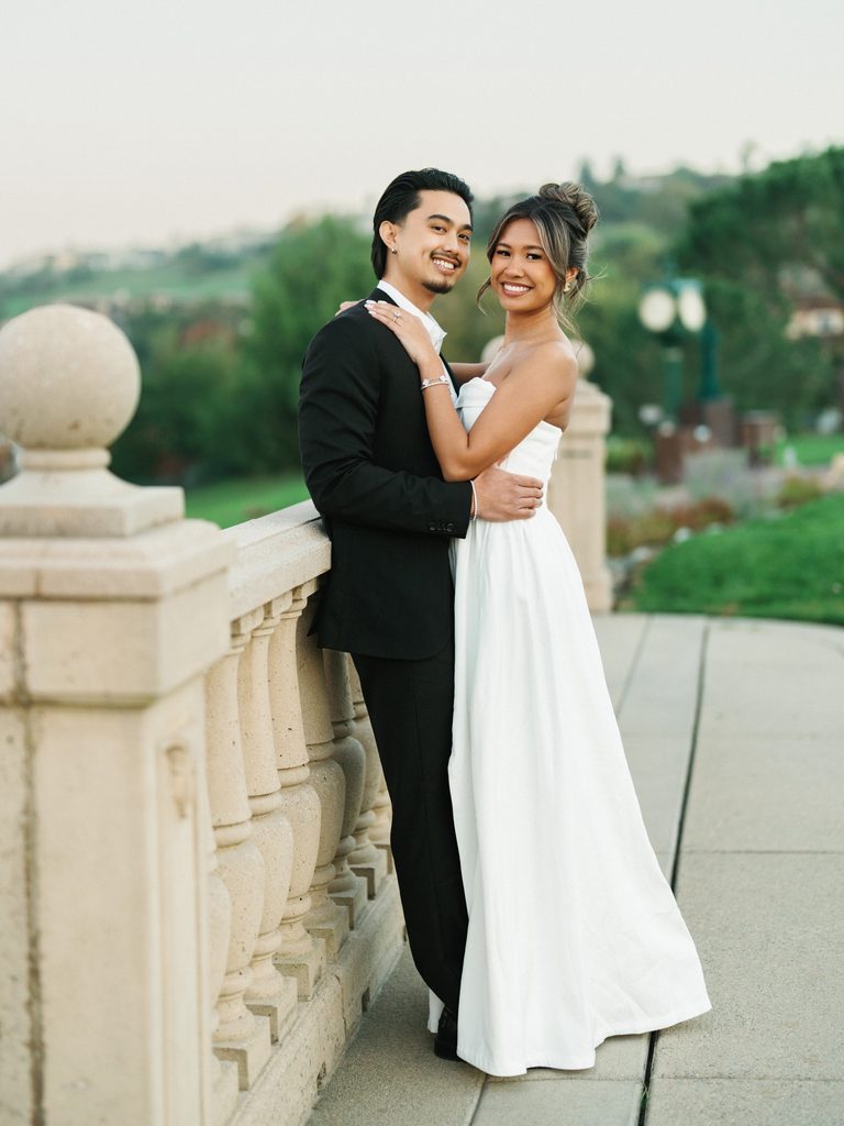 Chic bridal couple posing outdoor near a classic stone balustrade showing a bride in a strapless white gown and a groom in a black suit