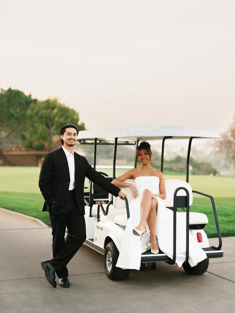 Stylish groom leaning against a golf cart while his beautiful bride sits inside showcasing her white block heels and elegant strapless dress