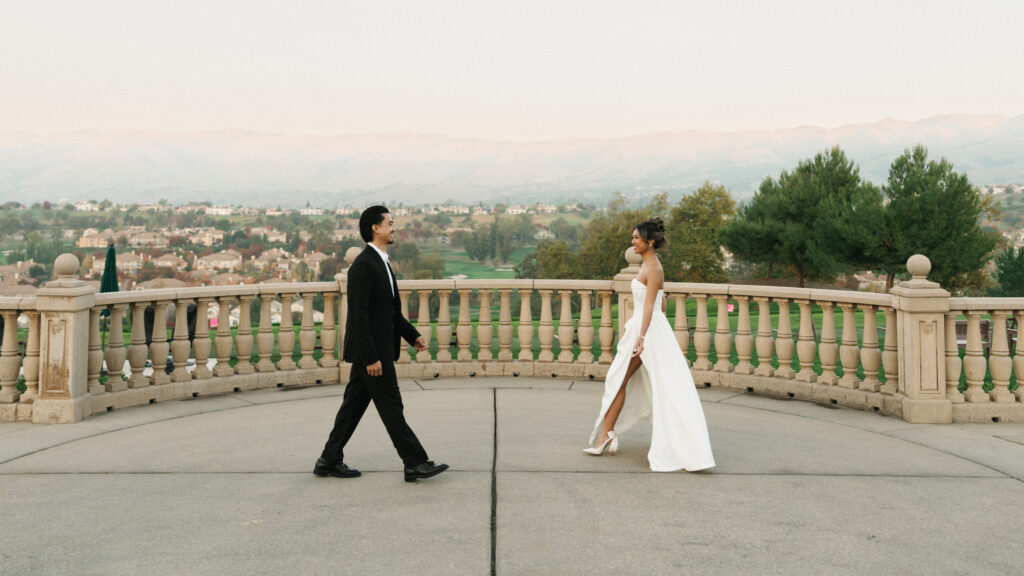 Stunning wide dynamic portrait of a bride and groom walking toward each other on a massive stone balcony with a classic balustrade overlooking beautiful mountains and valley hills