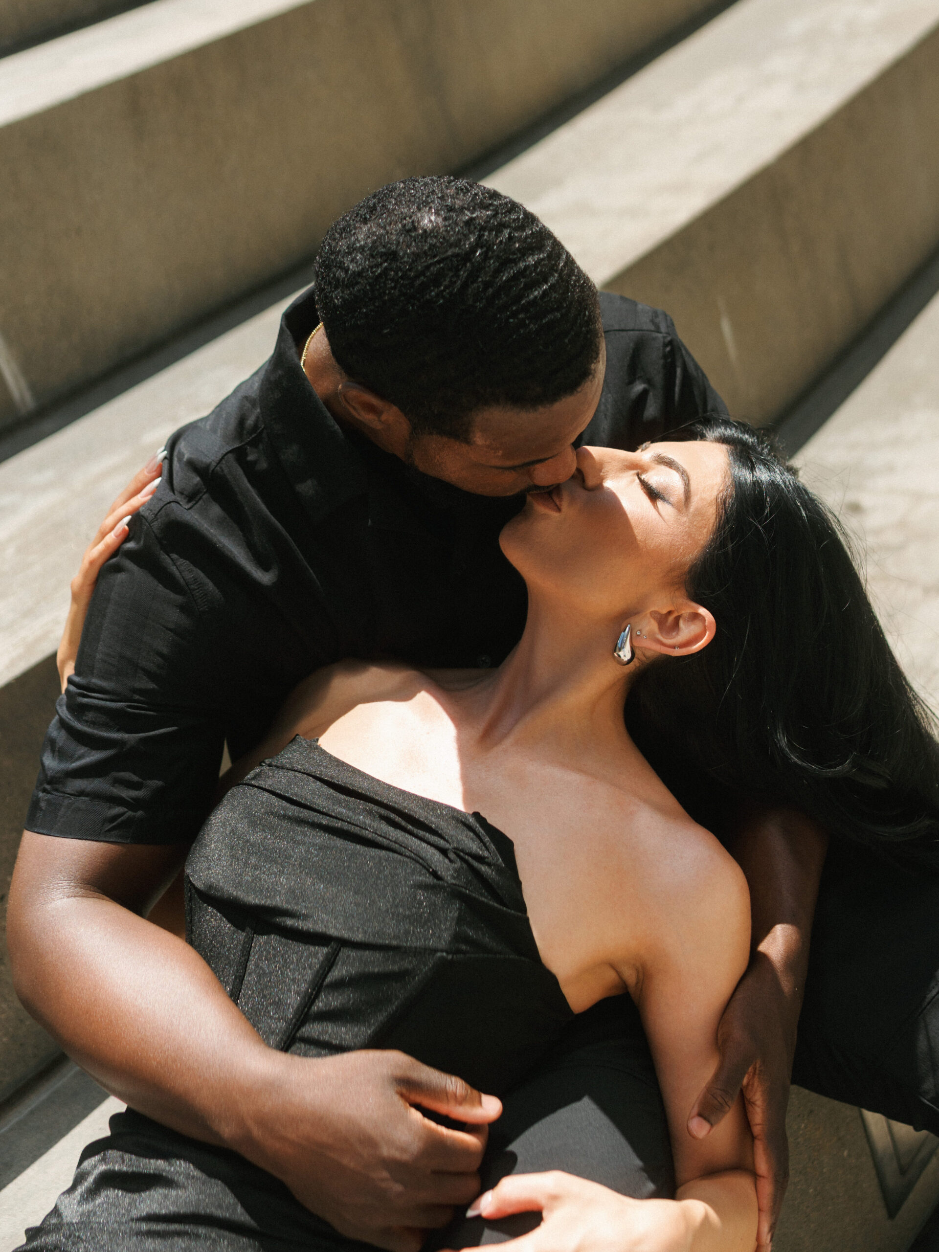 Man in black shirt kissing a woman in a strapless corset dress on concrete steps, showing monochrome engagement photo outfits.