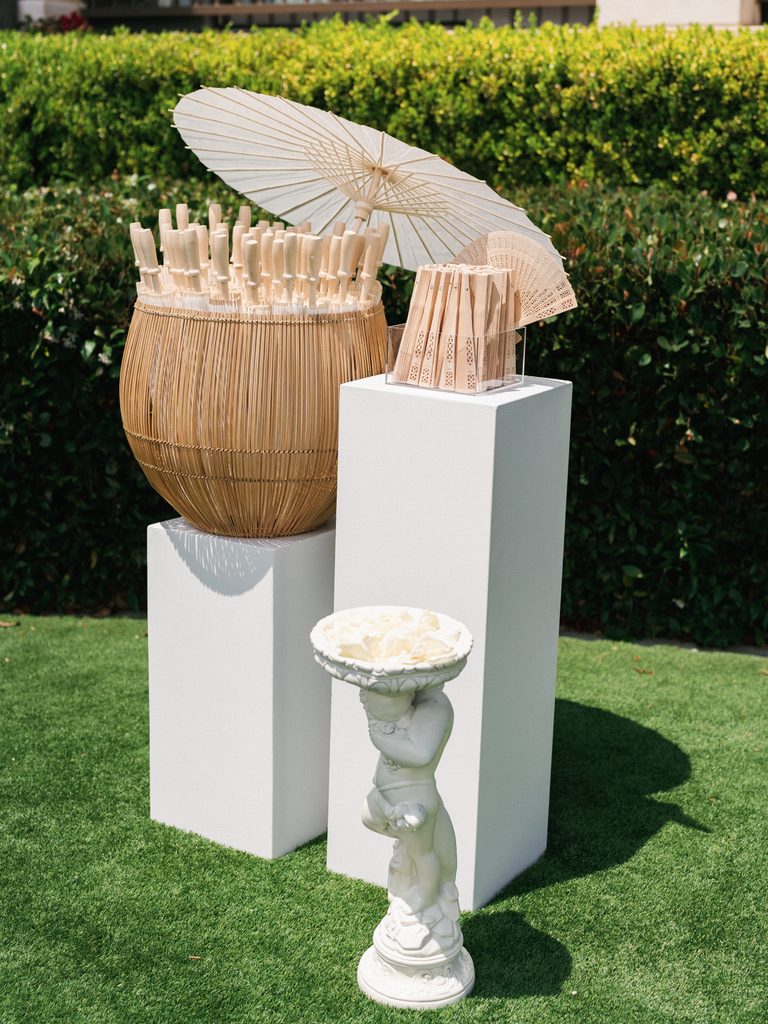 Upscale wedding ceremony photos showcasing a guest welcome station with wooden hand fans and white paper parasols on a lawn