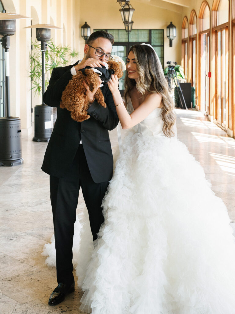 Bride and groom share a joyful moment with their small brown dog wearing a custom tuxedo inside a luxury venue corridor