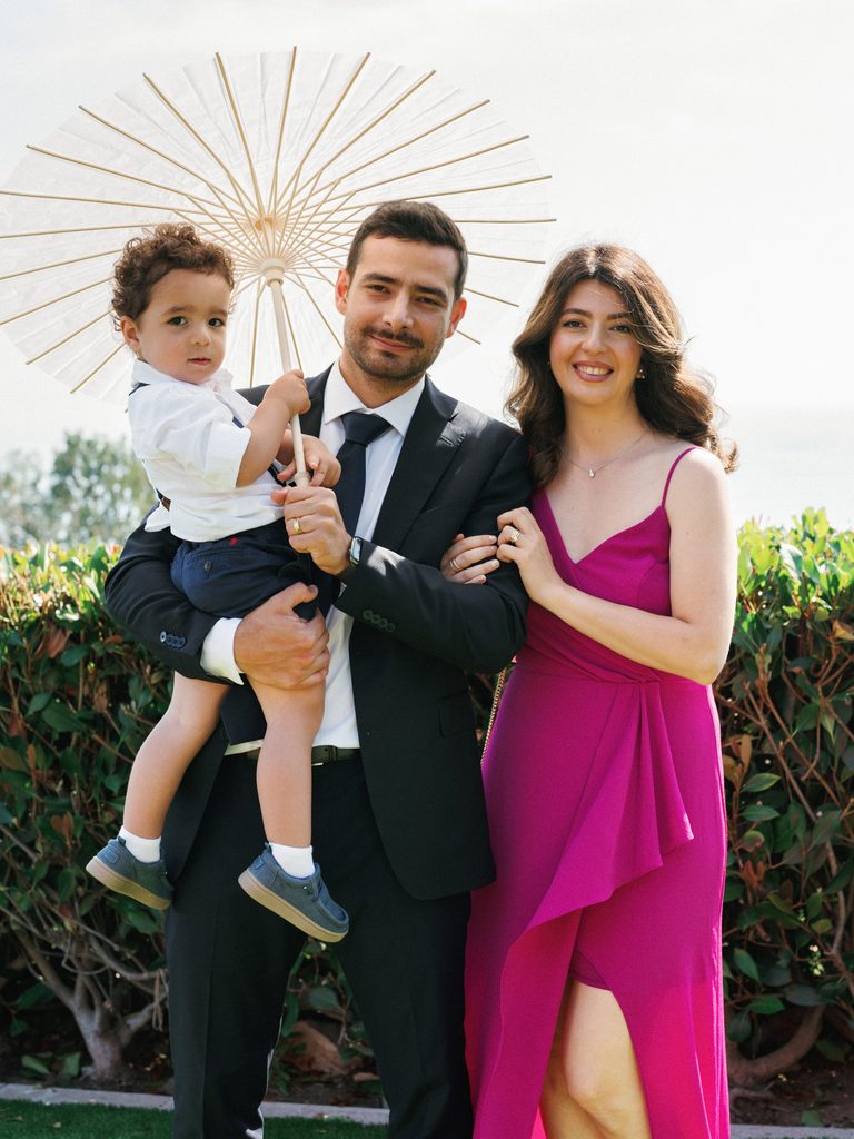 Elegant wedding guests smile with a toddler holding a white paper parasol during an outdoor coastal celebration