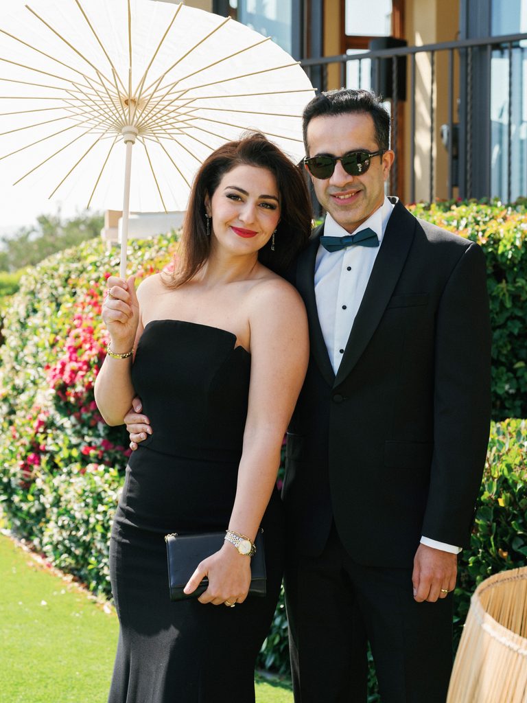 Stylish couple in a formal black dress and classic tuxedo poses under a sunny sky while holding a parasol