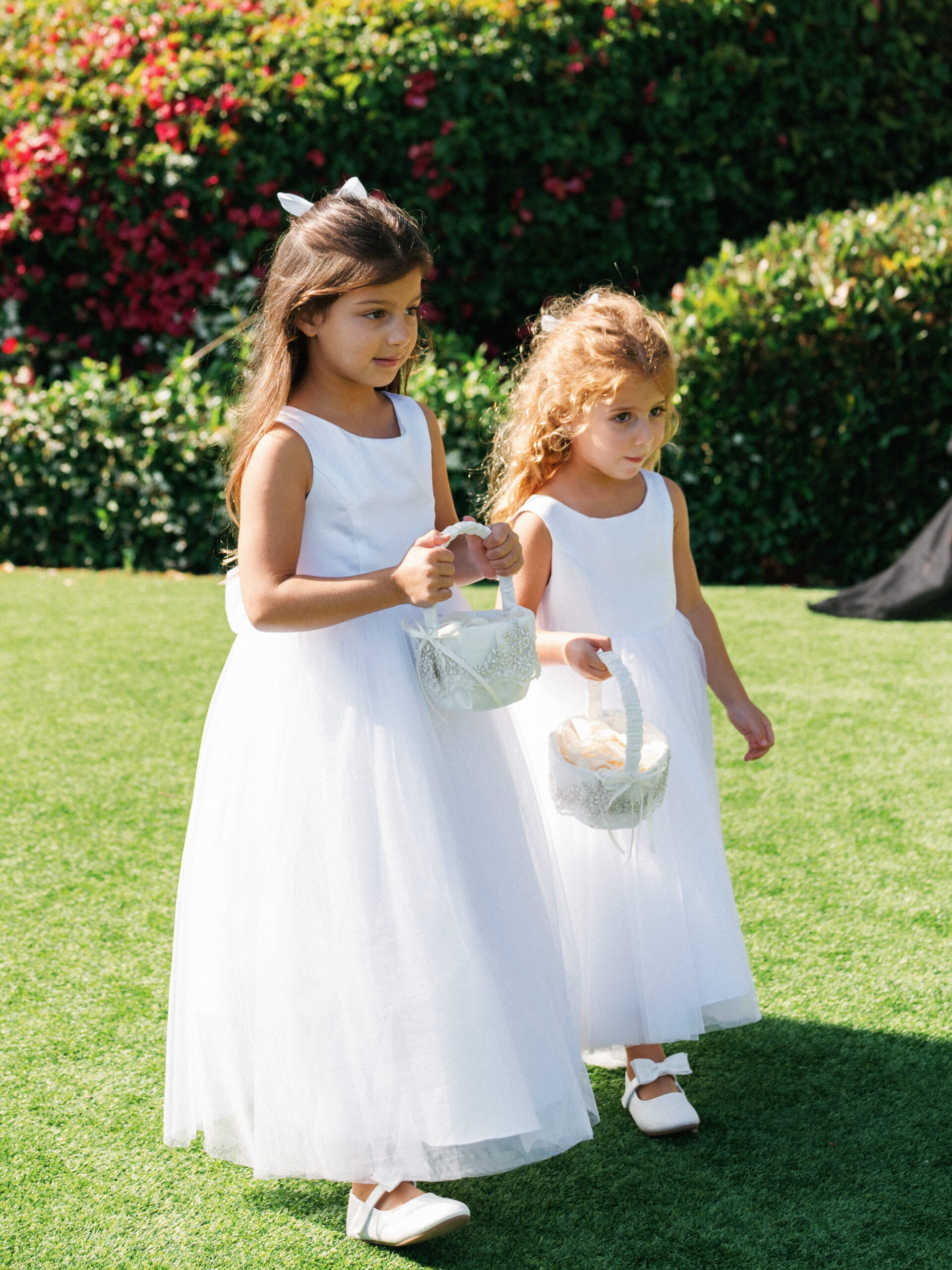 Two adorable flower girls in white dresses carry woven baskets during an outdoor ceremony