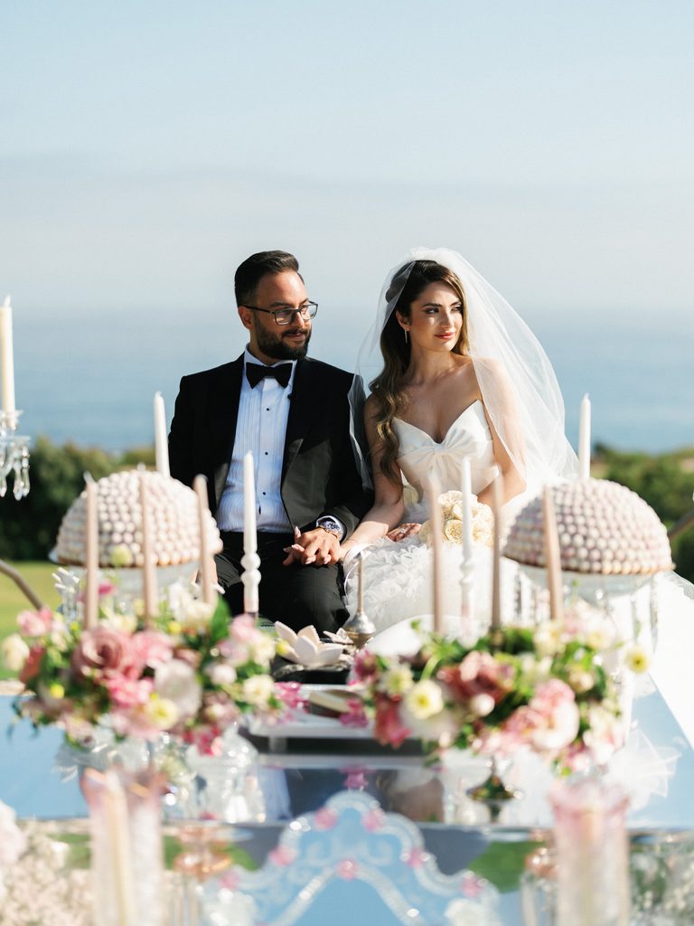 Bride and groom hold hands behind a mirrored altar table with floral arrangements overlooking the ocean