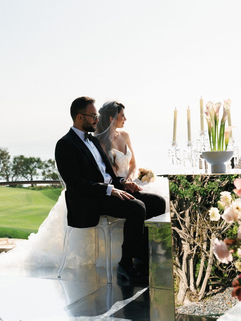 Newlyweds sit gracefully on acrylic chairs during an outdoor ceremony under the bright sun