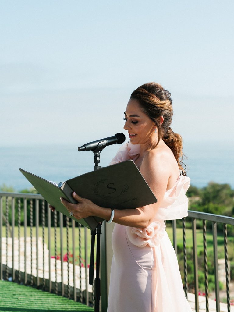 Wedding officiant reads from a monogrammed book into a microphone during a sunny outdoor ceremony