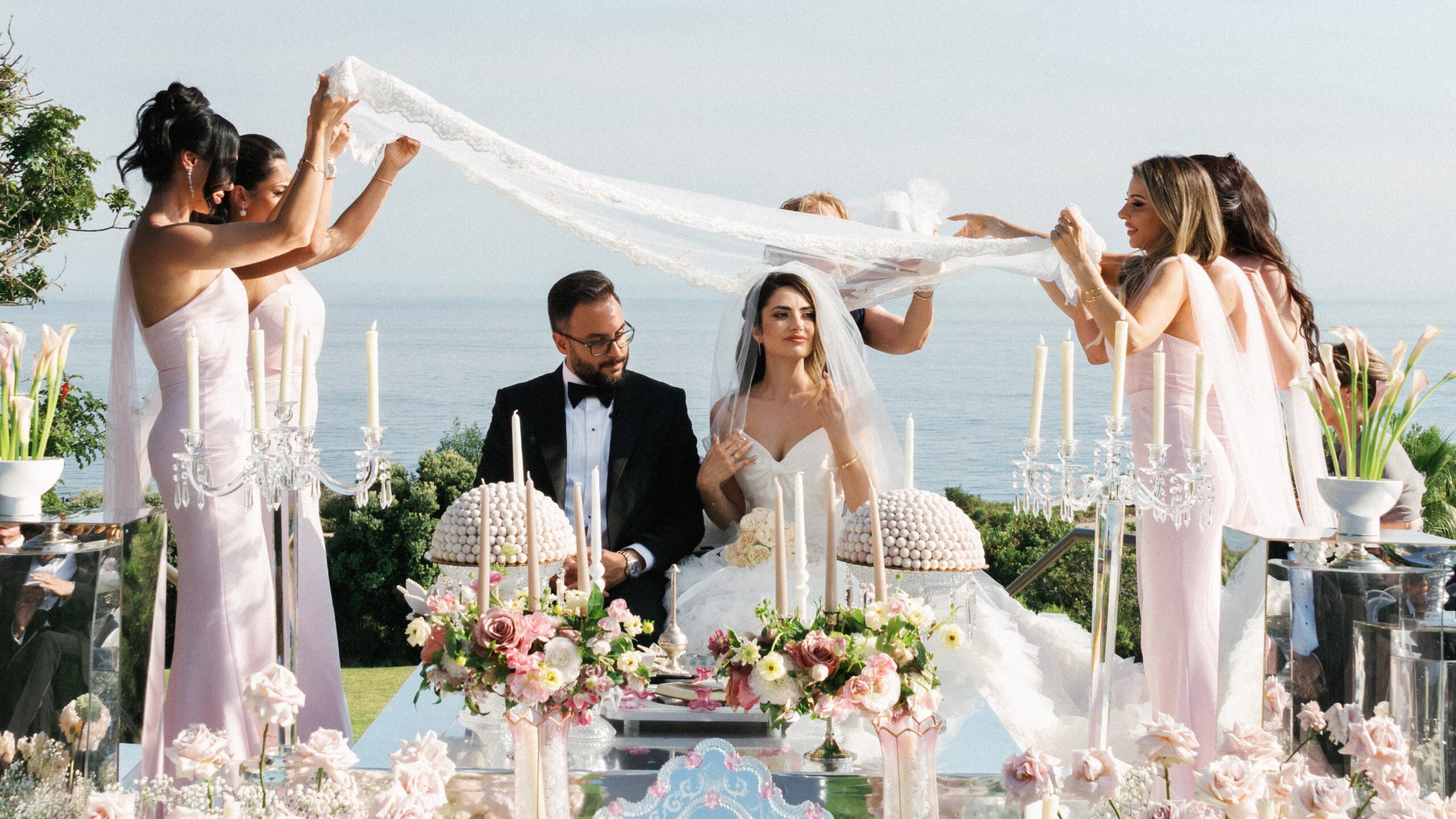 Bridesmaids hold a delicate lace veil over the bride and groom during a traditional Persian Sofreh Aghd ritual overlooking the ocean
