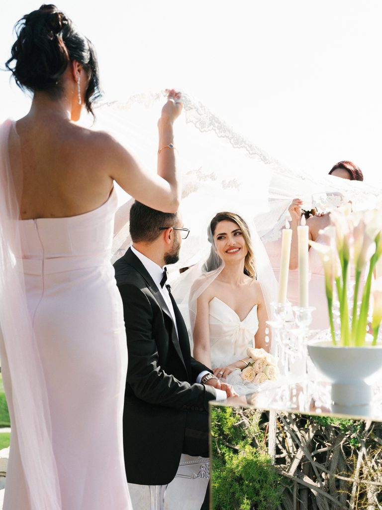 Bridesmaids hold a delicate lace veil over the seated couple during a traditional Persian wedding ritual