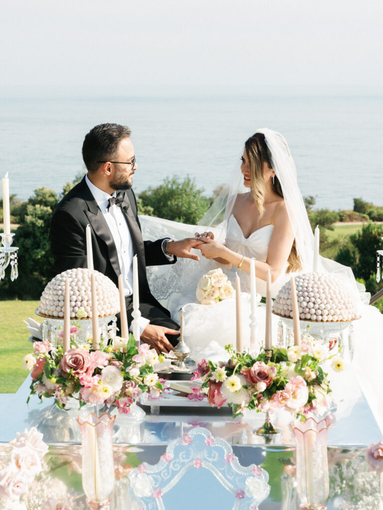 Bride smiles while placing a wedding ring on the grooms finger at their luxury outdoor Sofreh Aghd altar