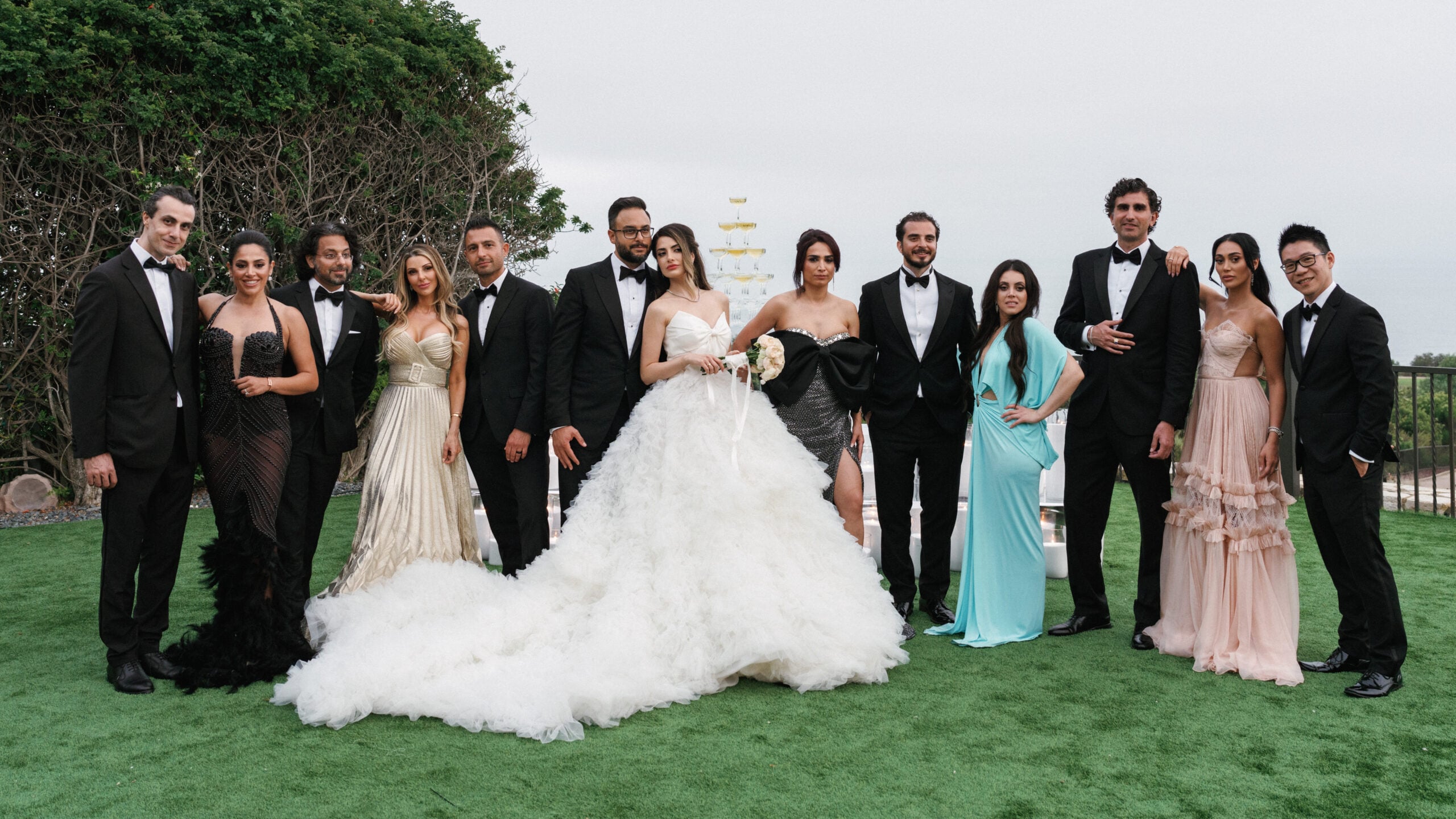 Newlyweds pose with their stylishly dressed wedding party and close friends on a lawn overlooking the ocean