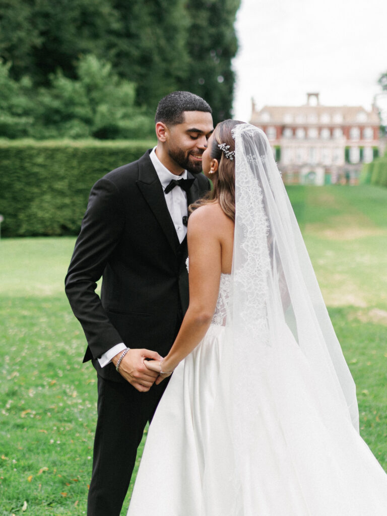 Sophisticated groom holding his brides hand and lean in for a kiss on a vast green lawn with a luxury mansion blurred in the background
