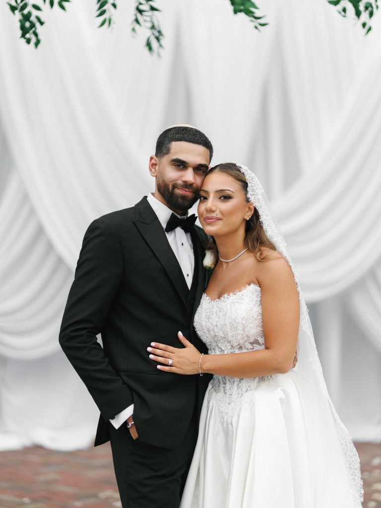 Elegant jewish bride and groom smiling for a portrait in front of a white drapery chuppah altar setup with green foliage
