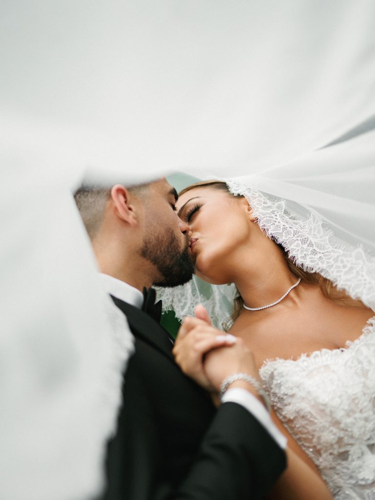 Cinematic close up of a couple kissing under a flowing white lace veil showing an elegant diamond necklace and lace corset gown detail