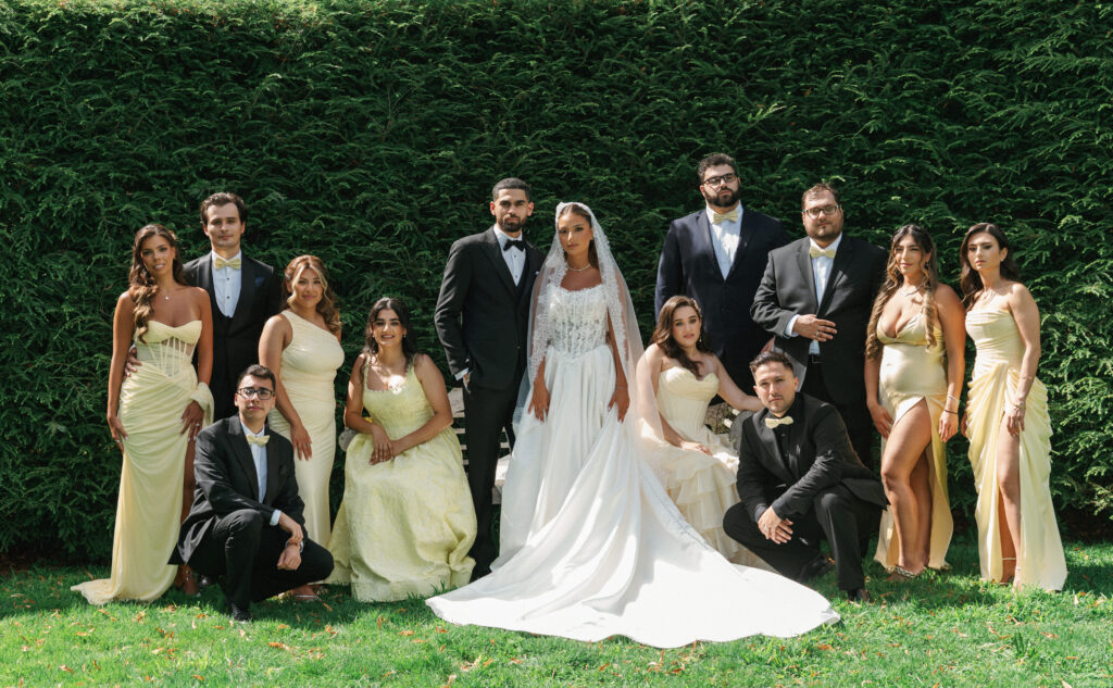Editorial wedding party posing outdoor in front of a green hedge with bridesmaids in elegant pastel yellow dresses and groomsmen in classic black tuxedos