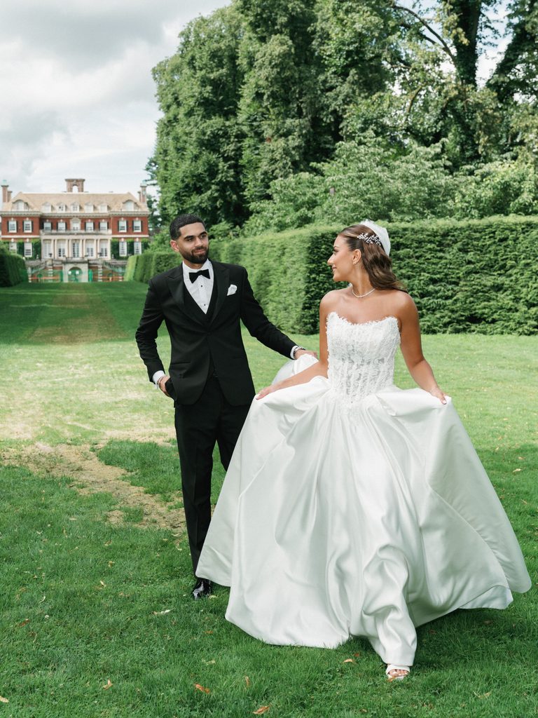 Stunning bride lifting her voluminous satin ballgown dress walking next to her groom near a historic estate manor architecture