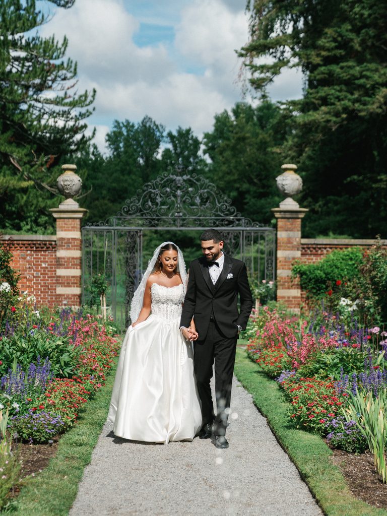 Breathtaking bridal couple walking hand in hand along a stone path in a lush blooming garden with a historic wrought iron gate