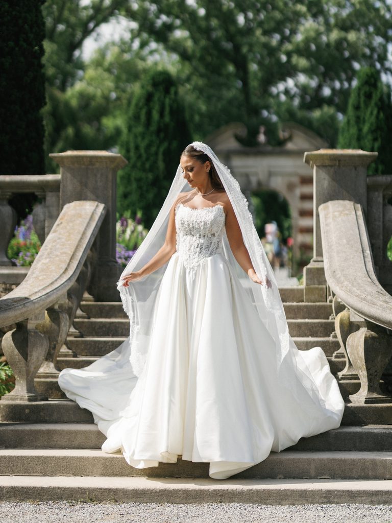 Breathtaking bride walking down historic stone stairs in a luxury lace corset gown with a long embroidered veil