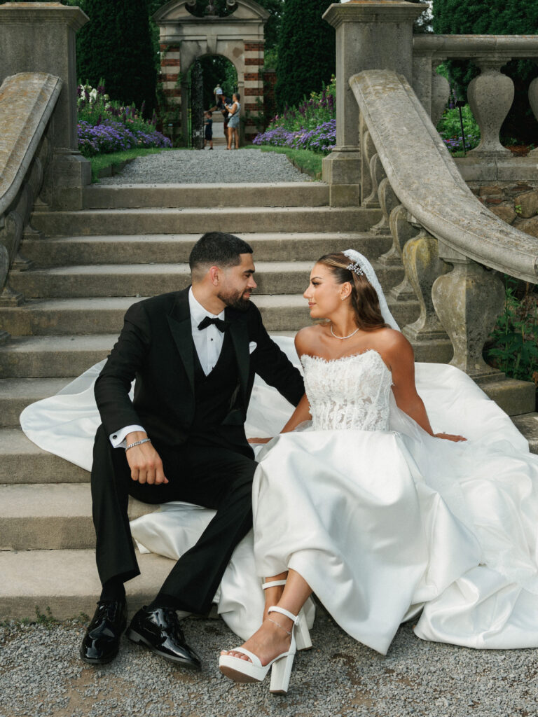 Chic bridal couple sitting on stone garden stairs looking at each other showcasing white block heel platform sandals and a classic three piece black tuxedo