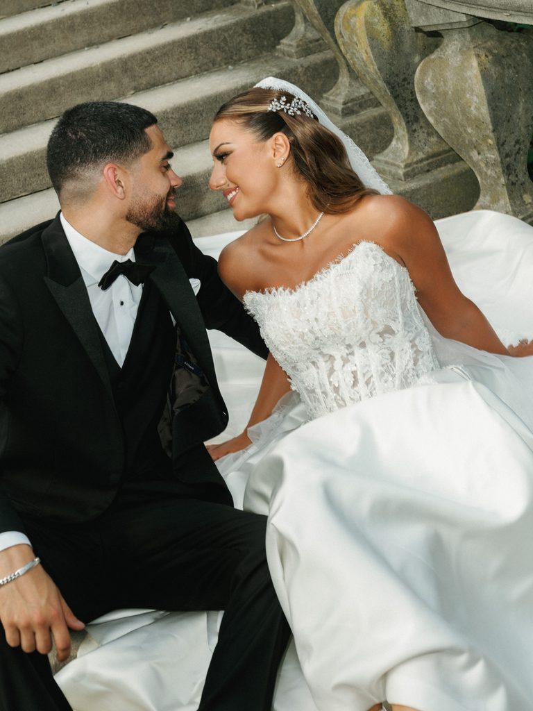 Intimate wedding couple portrait on stone steps showing a smiling bride and groom looking at each other featuring an ornate lace corset dress and diamond necklace
