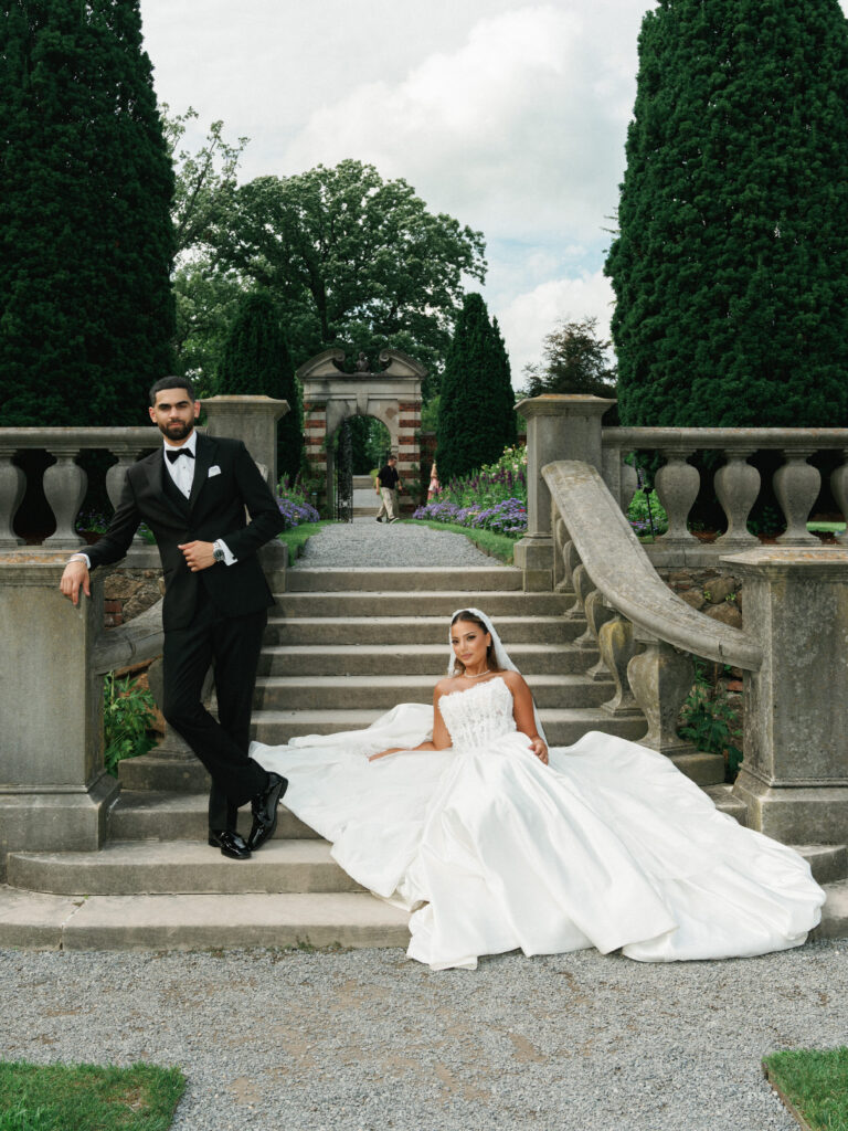 Editorial wedding portrait on historic estate steps featuring a bride sitting with a massive spread out ballgown train and a groom standing next to stone balustrade