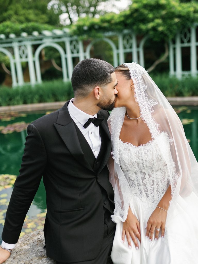 Cinematic close up portrait of newlyweds kissing next to a scenic garden pond showing an elegant lace trim veil and modern black tuxedo