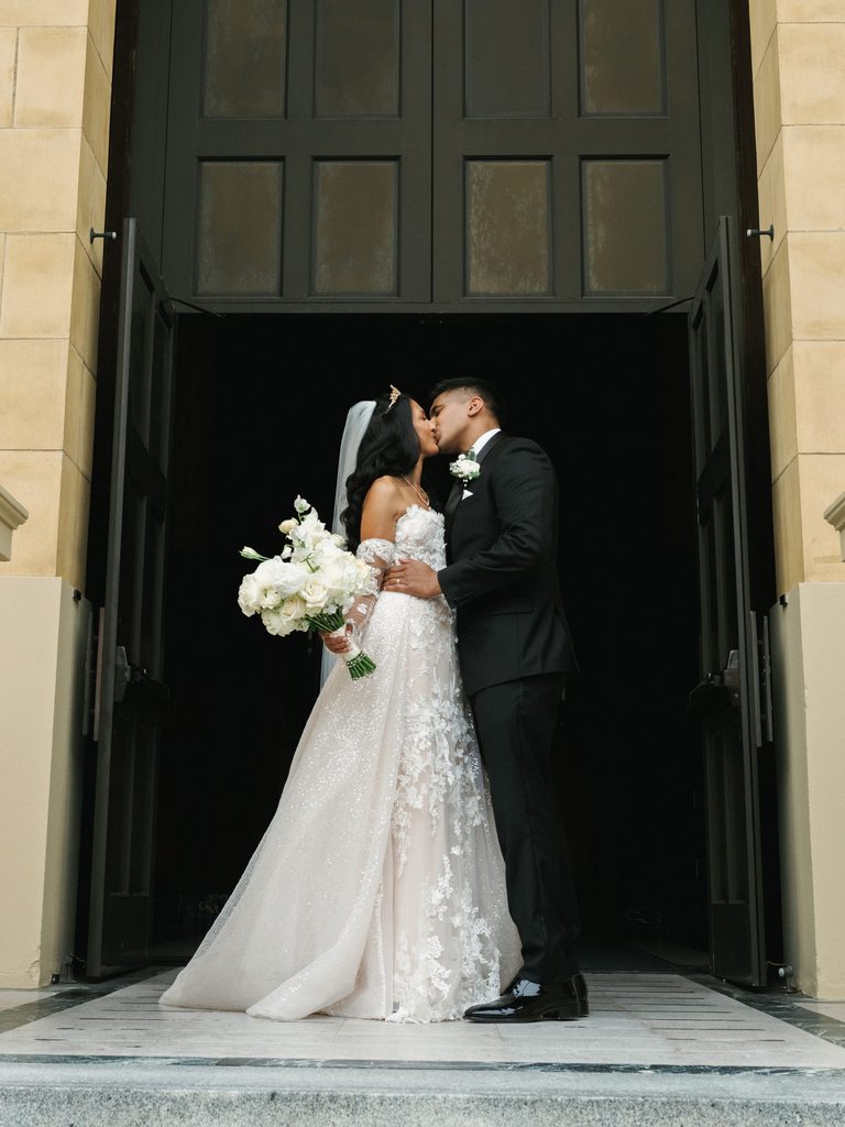 Cinematic newlyweds kiss in front of grand dark church doors highlighting a voluminous sparkling bridal gown and lush white rose bouquet