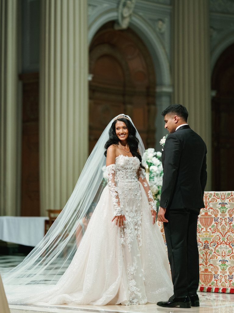 Radiant bride in a strapless ballgown smiling at her groom while standing in front of majestic church columns during their wedding ceremony