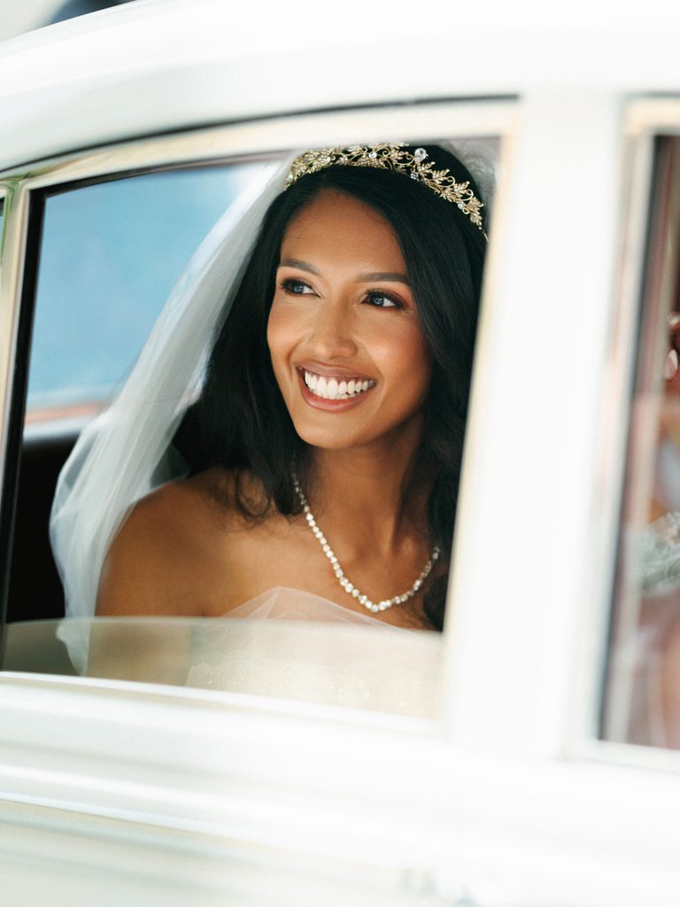 Gorgeous bride smiling looking through a vintage wedding car window showcasing her elegant gold tiara and delicate pearl necklace
