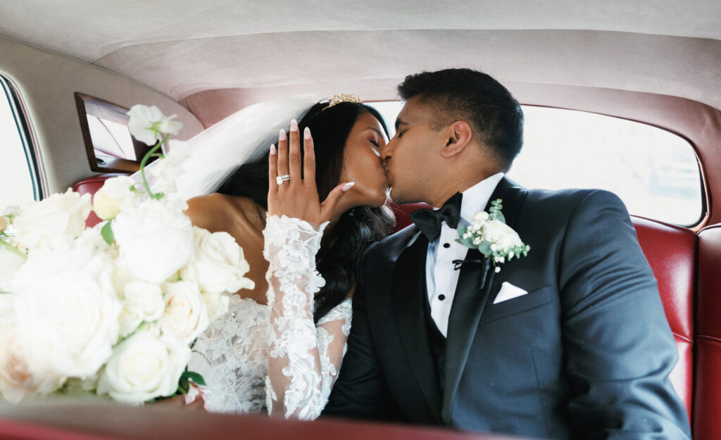 Intimate newlyweds kissing inside a vintage luxury getaway car with red leather interior while the bride shows her elegant diamond ring
