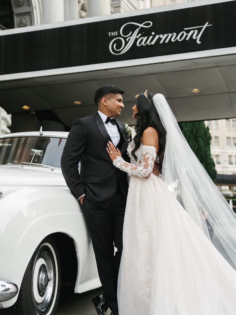 Chic newlyweds posing next to a classic white getaway car under the iconic fairmont hotel awning for a luxury city wedding portrait session