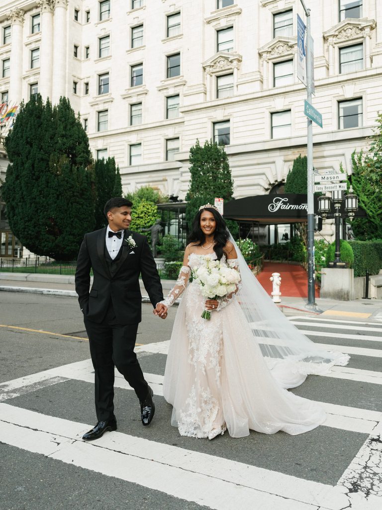 Modern luxury newlyweds smiling and crossing an urban road near the famous fairmont hotel entrance with a lush white bouquet
