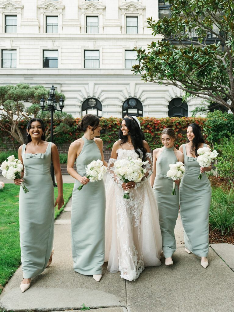 Joyful bride walking and laughing outdoor with bridesmaids wearing elegant long sage green dresses and holding lush white bouquets