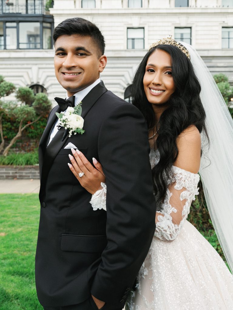 Chic bridal couple posing outdoor on a manicured green lawn with a bride gently leaning on her grooms shoulder showing a luxury diamond ring
