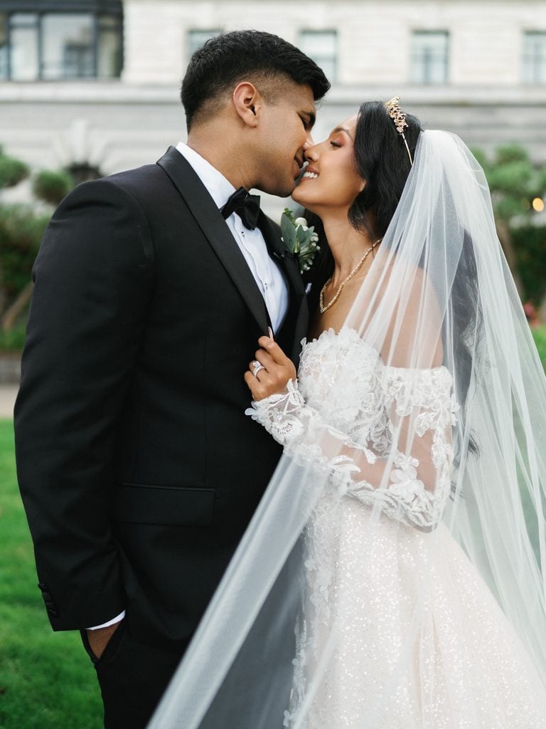 Cinematic wedding day couple portrait of a groom kissing his bride wearing a luxury off the shoulder lace gown with an elegant gold tiara and flowing veil