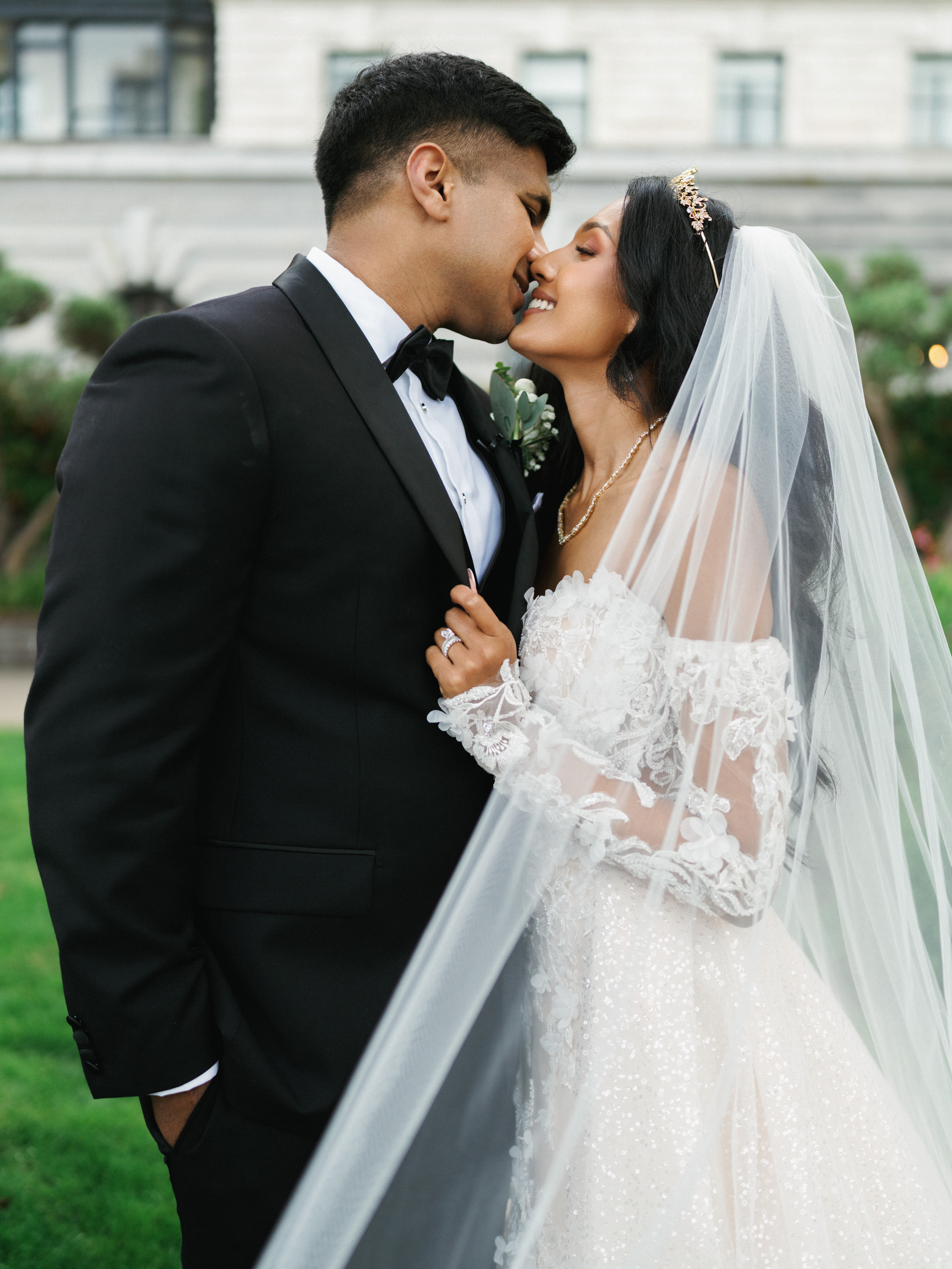 Cinematic wedding day couple portrait of a groom kissing his bride wearing a luxury off the shoulder lace gown with an elegant gold tiara and flowing veil