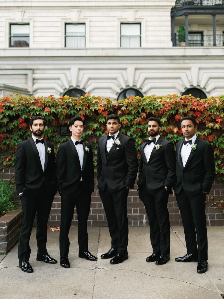 Sophisticated groomsmen line up portrait with a groom posing in classic sharp black tuxedos against a lush ivy brick wall