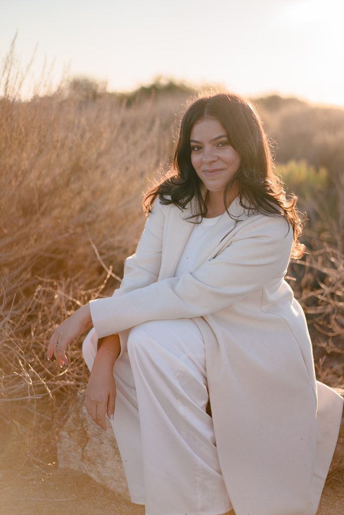 Future bride sitting on a rock in a cream-colored pantsuit amidst dry grass at sunset.